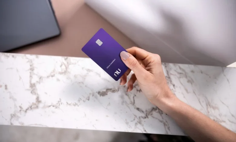 Cartão Nubank ultravioleta sendo segurado por uma pessoa sobre uma mesa de mármore, em um cenário minimalista.