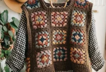 Imagem de um colete de crochê estilo granny square com quadrados coloridos em tons de marrom, azul, laranja e rosa, pendurado em um cabide sobre um fundo claro.
