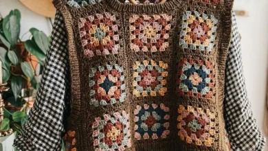 Imagem de um colete de crochê estilo granny square com quadrados coloridos em tons de marrom, azul, laranja e rosa, pendurado em um cabide sobre um fundo claro.