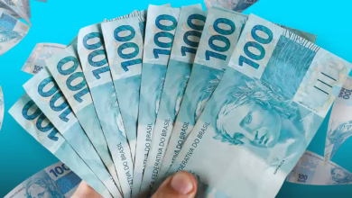 Imagem de várias cédulas de R$ 100, com foco em uma mão segurando uma pilha, simbolizando o pagamento do Bolsa Família e a consulta pelo Caixa Tem.