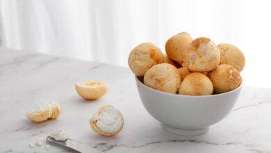 Imagem de uma tigela com pães de queijo frescos e macios ao lado de um pão de queijo aberto com recheio de queijo, sobre uma superfície de mármore
