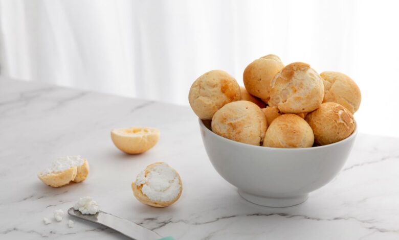 Imagem de uma tigela com pães de queijo frescos e macios ao lado de um pão de queijo aberto com recheio de queijo, sobre uma superfície de mármore