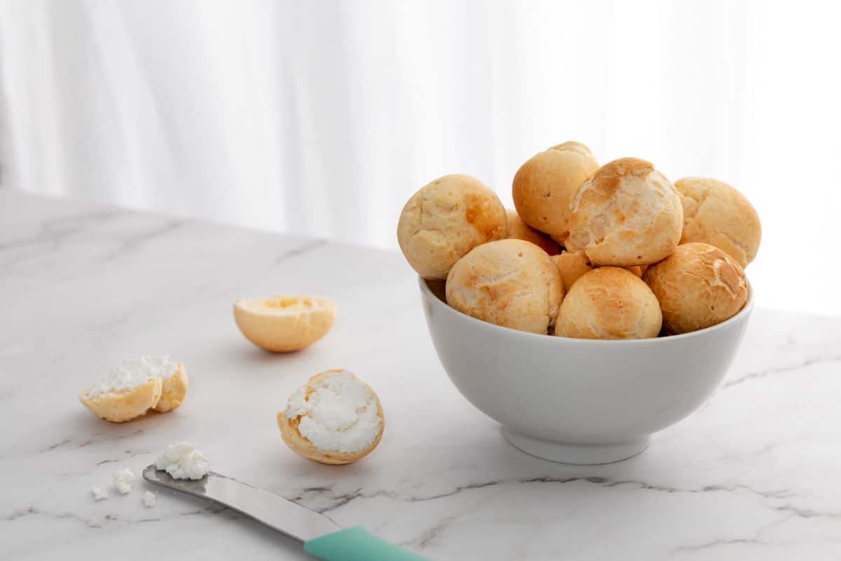 Imagem de uma tigela com pães de queijo frescos e macios ao lado de um pão de queijo aberto com recheio de queijo, sobre uma superfície de mármore