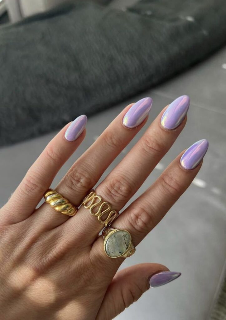 Mão com unhas amendoadas pintadas com esmalte perolado em tom lavanda, usando anéis dourados.