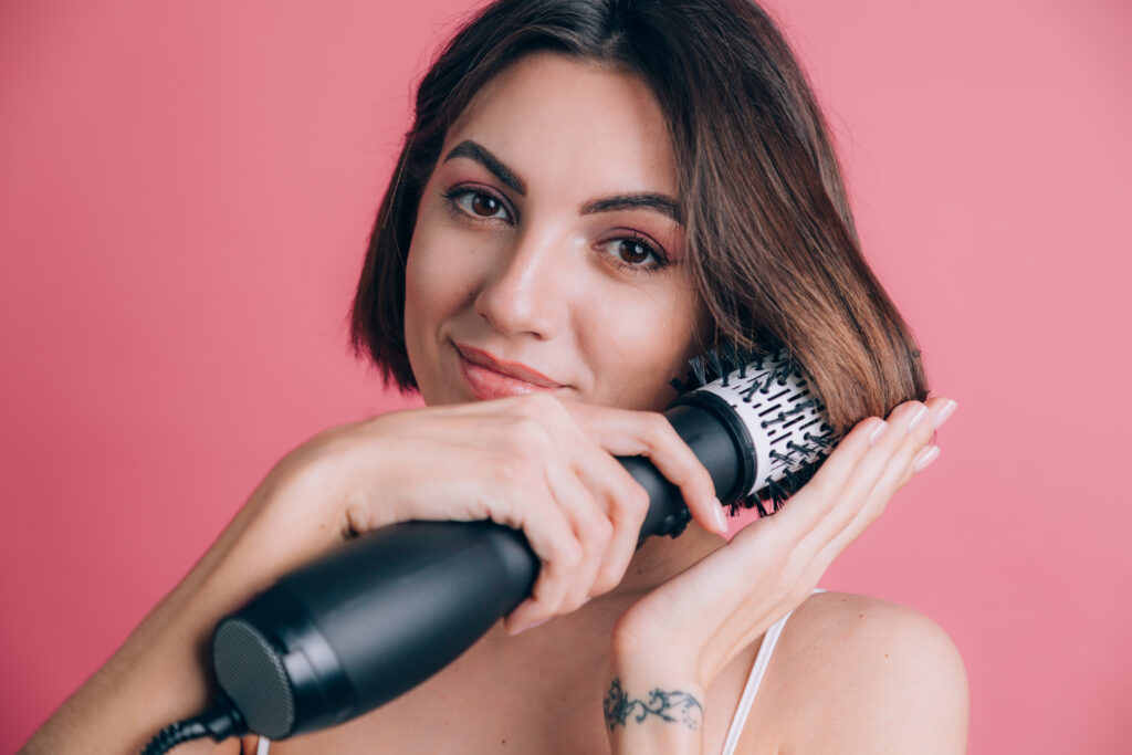 Mulher usando uma escova modeladora para secar e pentear o cabelo, com um sorriso leve.