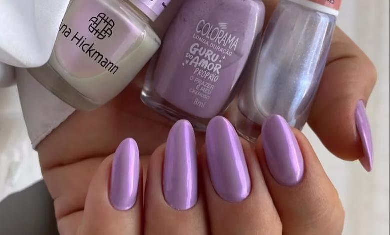 Unhas esmaltadas em tom lilás espelhado com três vidrinhos de esmalte ao fundo