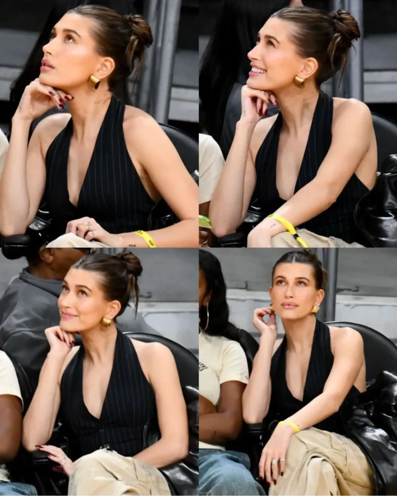 Hailey Bieber com cabelo sleek em evento, usando blusa preta e calça clara, observando atentamente.