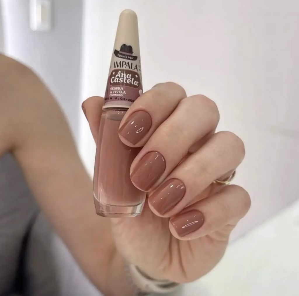 Imagem de unhas pintadas com esmalte marrom claro, com a embalagem do esmalte visível, destacando a tonalidade e o brilho.
