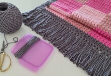 Acessórios e materiais para crochê, incluindo um novelo de lã cinza, tesoura e uma peça de crochê rosa, prontos para a confecção de um tapete.