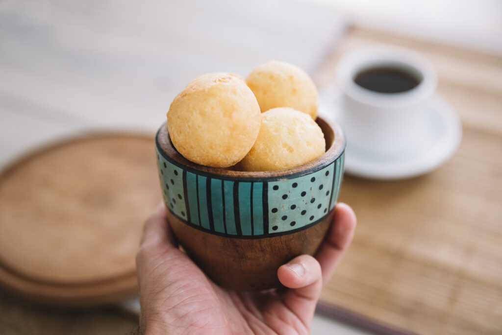 Pessoa segurando uma tigela artesanal com pães de queijo mineiro frescos ao lado de uma xícara de café.