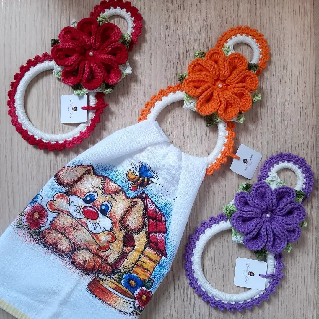 Porta-pano de prato em crochê com flores coloridas em vermelho, laranja e roxo, combinados com toalha estampada de cachorro e flores.