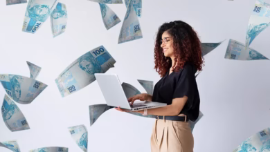 Mulher com laptop, sorrindo enquanto observa cédulas de 100 reais caindo, representando o rendimento da conta Nubank em agosto.