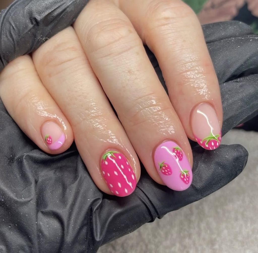 Unhas curtas decoradas com desenhos de morangos em fundo rosa.