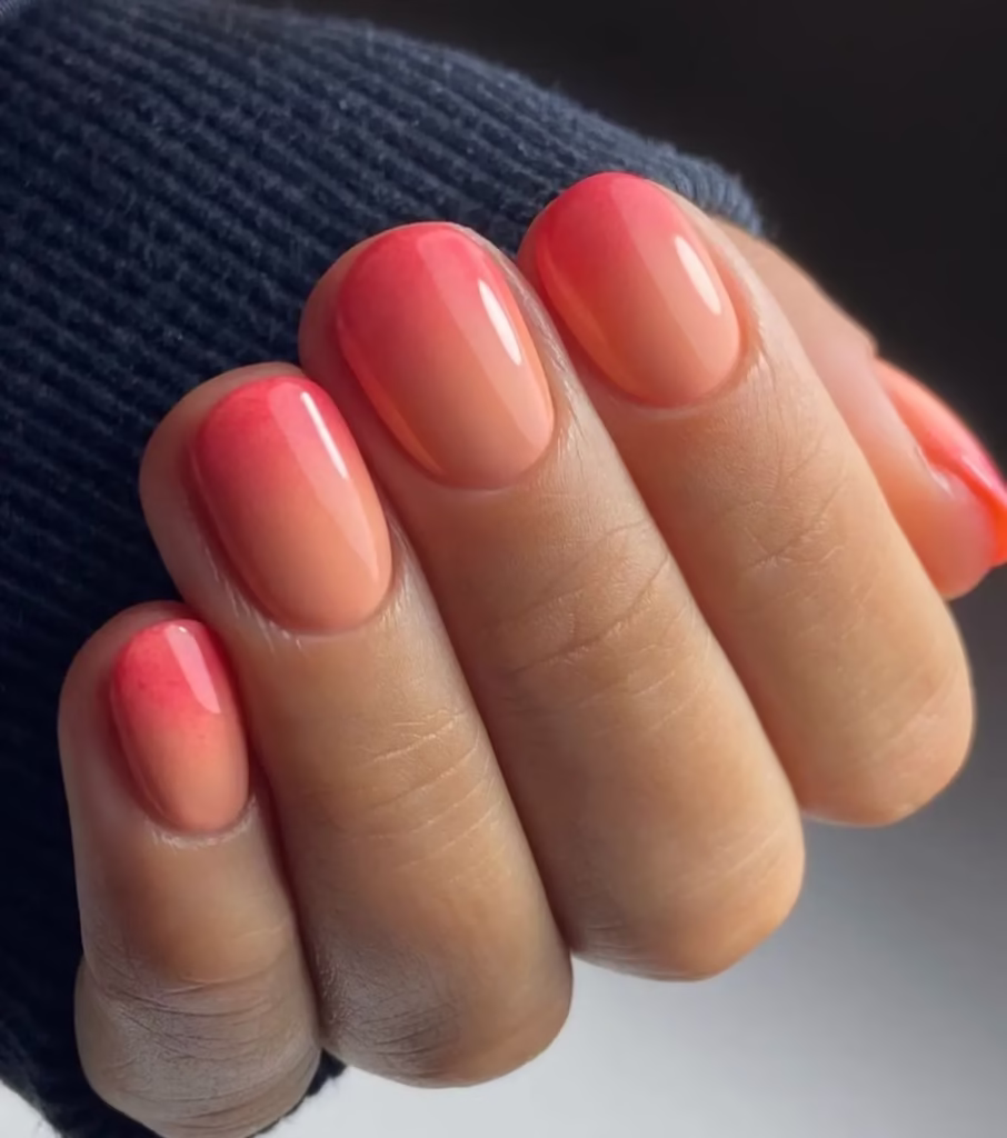 Unhas curtas com efeito degradê em tons de coral e rosa.