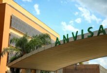 Fachada da sede da Anvisa, Agência Nacional de Vigilância Sanitária, com seu nome em letras verdes grandes e palmeiras ao redor.