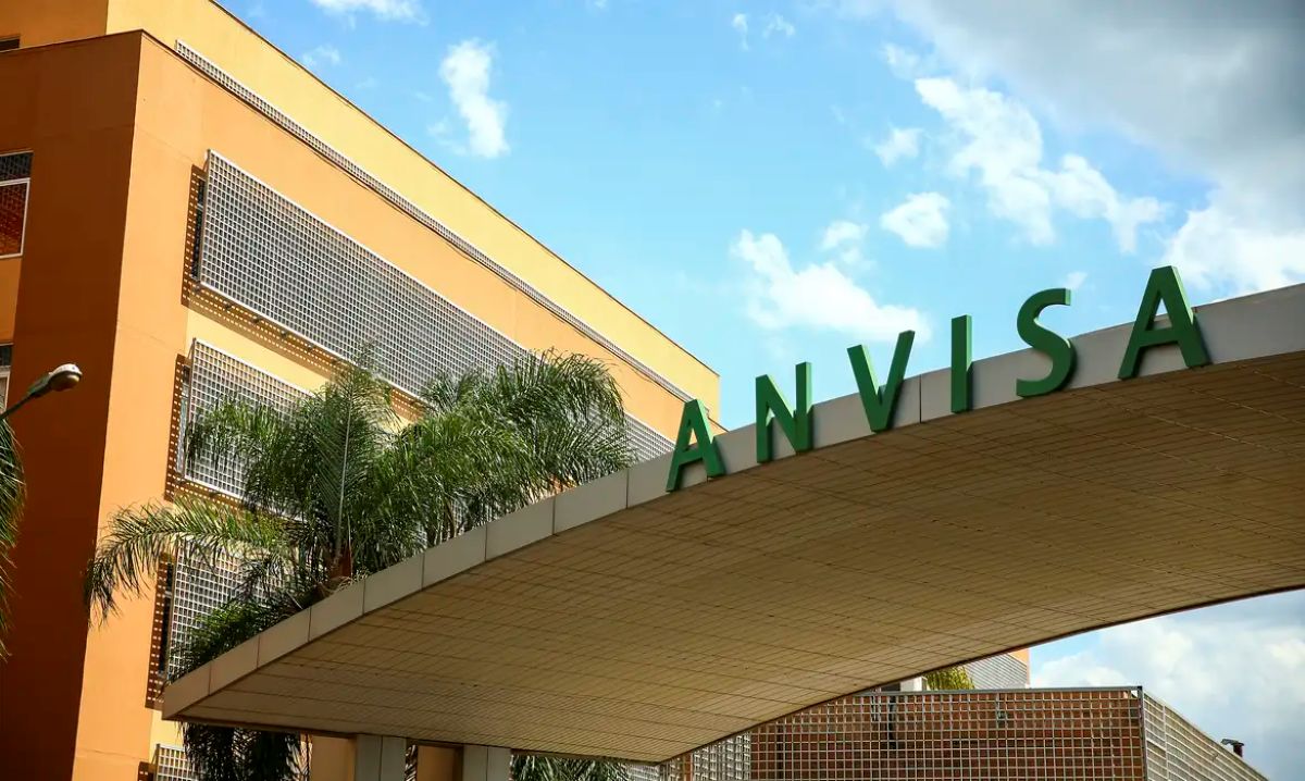 Fachada da sede da Anvisa, Agência Nacional de Vigilância Sanitária, com seu nome em letras verdes grandes e palmeiras ao redor.