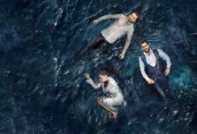 Três atores de novela turca flutuando em águas escuras. Os homens vestem camisas sociais e a mulher, um vestido branco.