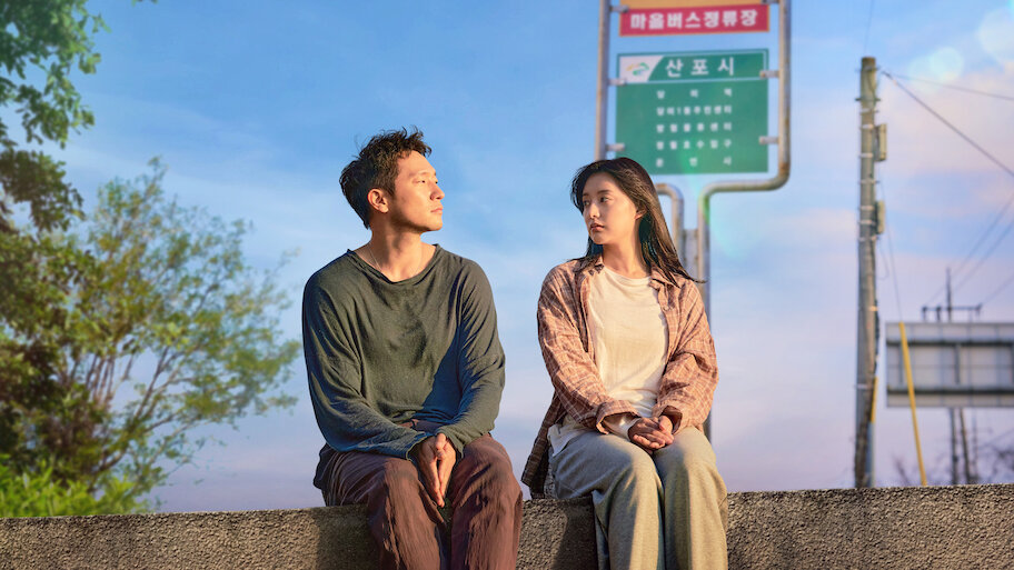 Os dois protagonistas do dorama 'Meu Diário para a Liberdade' sentados em uma mureta, olhando um para o outro em uma cena noturna, com uma placa de ponto de ônibus ao fundo.