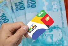 Cartão do programa Bolsa Família em primeiro plano, com cédulas de 100 reais ao fundo, destacando o pagamento do benefício.