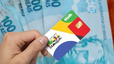 Cartão do programa Bolsa Família em primeiro plano, com cédulas de 100 reais ao fundo, destacando o pagamento do benefício.