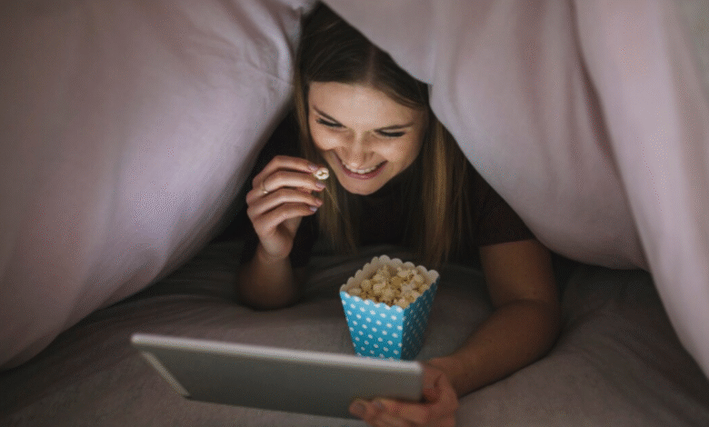 Mulher sorrindo, escondida sob um edredom, assistindo a algo em seu tablet enquanto come pipoca.