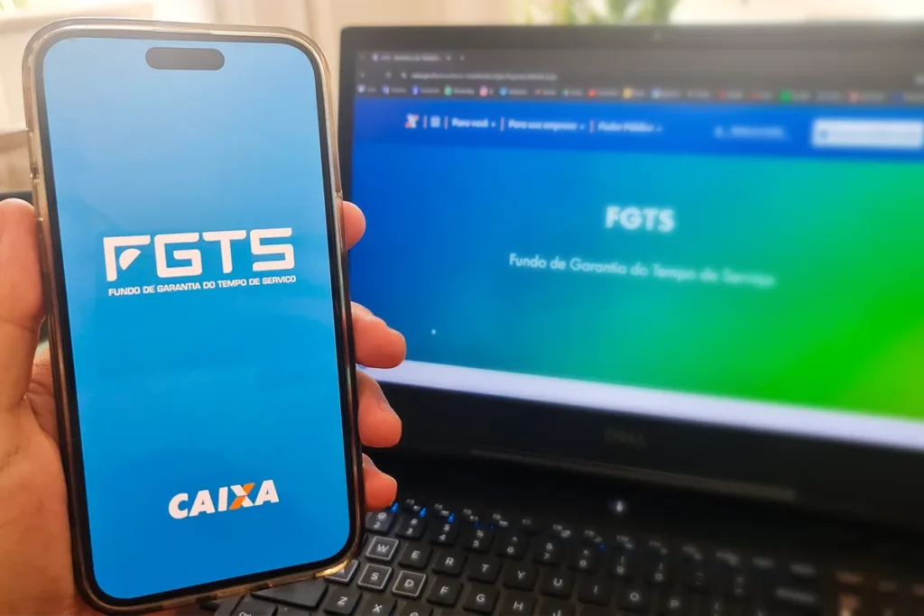 Uma mão segura um maço de notas de R$ 100 ao lado de um smartphone que exibe o logo do FGTS. A imagem simboliza a regularização financeira de débitos relacionados ao Fundo de Garantia.