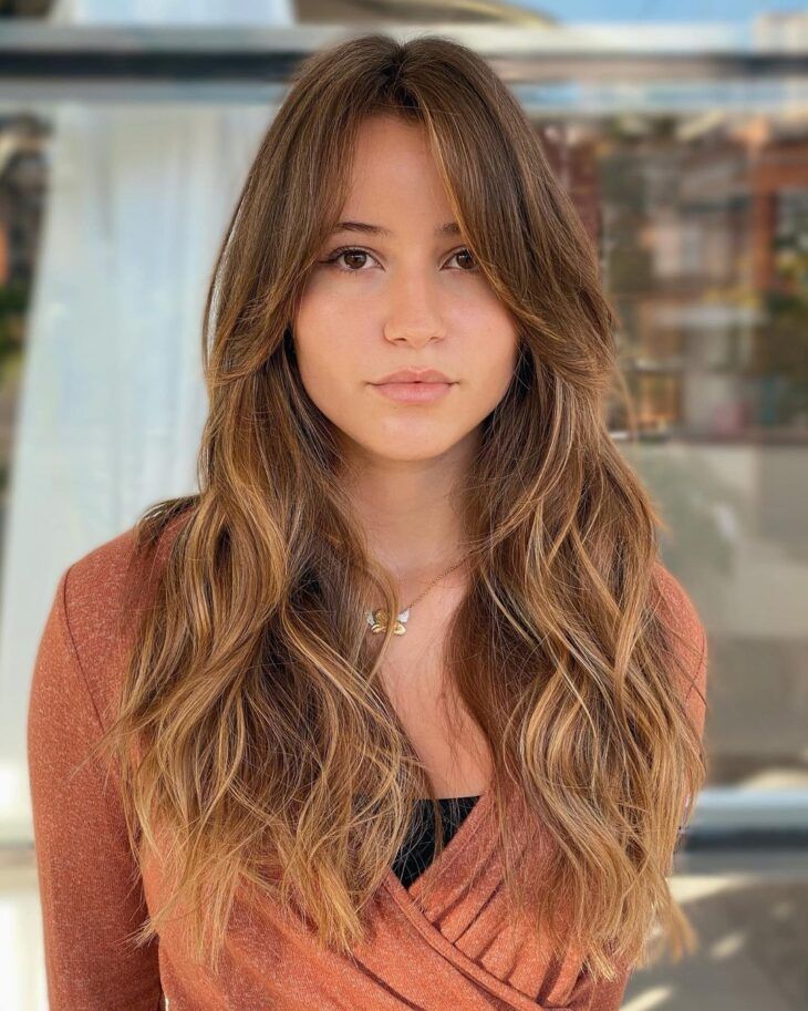 Mulher com cabelo castanho e mechas loiras, exibindo franja cortina longa e moderna.