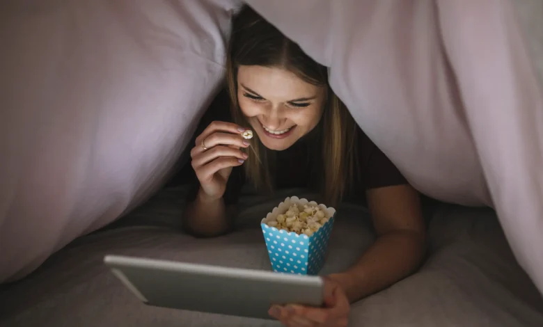 Pessoa assistindo a filmes na cama, comendo pipoca enquanto usa um tablet em um ambiente confortável
