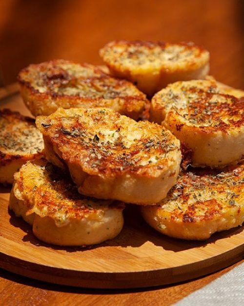 Fatias de rabanada salgada com queijo e orégano, dispostas em um prato de madeira. As fatias estão douradas e crocantes, prontas para serem servidas.