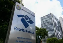 Placa da Receita Federal com logotipo e edifício ao fundo, sede do Ministério da Fazenda.