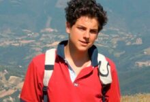 Jovem sorrindo, Carlo Acutis, em um campo verde com montanhas ao fundo, usando uma mochila e uma camisa polo vermelha.