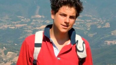 Jovem sorrindo, Carlo Acutis, em um campo verde com montanhas ao fundo, usando uma mochila e uma camisa polo vermelha.