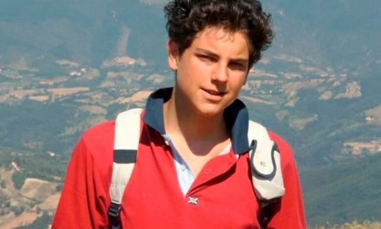 Jovem sorrindo, Carlo Acutis, em um campo verde com montanhas ao fundo, usando uma mochila e uma camisa polo vermelha.