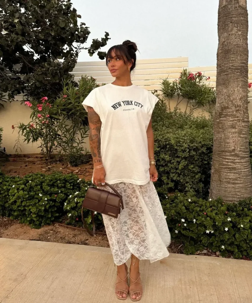 Mulher usando camiseta oversized branca com estampa 'New York City' e saia de renda transparente branca em ambiente ao ar livre.