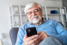 Homem sênior sorrindo enquanto ouve ou lê algo no celular com fones de ouvido, ilustrando o envio ou o recebimento de uma mensagem de início de semana motivacional pelo WhatsApp.