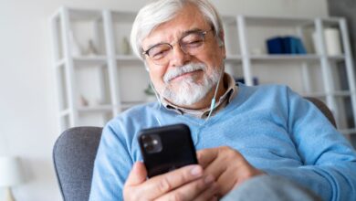 Homem sênior sorrindo enquanto ouve ou lê algo no celular com fones de ouvido, ilustrando o envio ou o recebimento de uma mensagem de início de semana motivacional pelo WhatsApp.