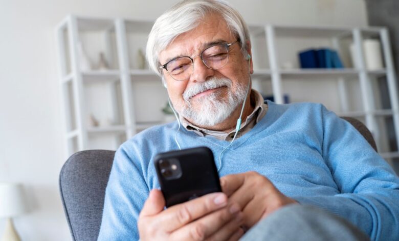 Homem sênior sorrindo enquanto ouve ou lê algo no celular com fones de ouvido, ilustrando o envio ou o recebimento de uma mensagem de início de semana motivacional pelo WhatsApp.