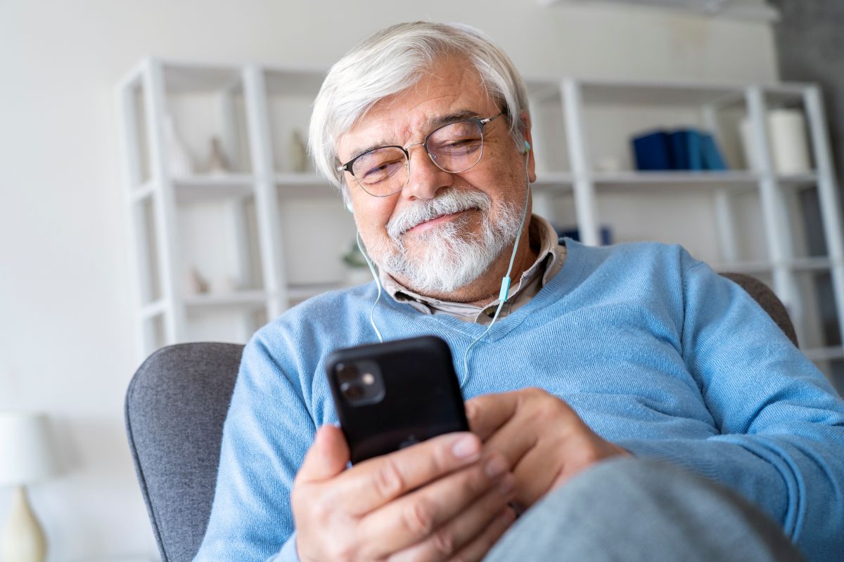 Homem sênior sorrindo enquanto ouve ou lê algo no celular com fones de ouvido, ilustrando o envio ou o recebimento de uma mensagem de início de semana motivacional pelo WhatsApp.