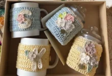 Copos de café com capas de crochê coloridas, com detalhes florais e iniciais, em caixa de presente.