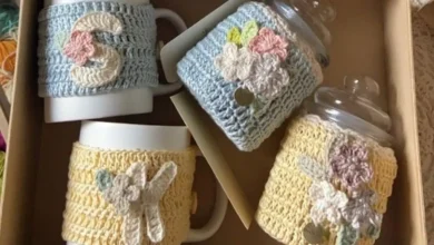 Copos de café com capas de crochê coloridas, com detalhes florais e iniciais, em caixa de presente.