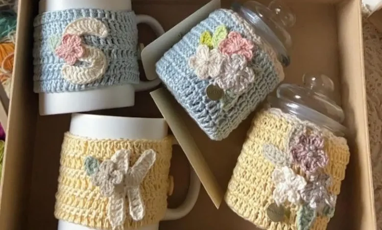 Copos de café com capas de crochê coloridas, com detalhes florais e iniciais, em caixa de presente.