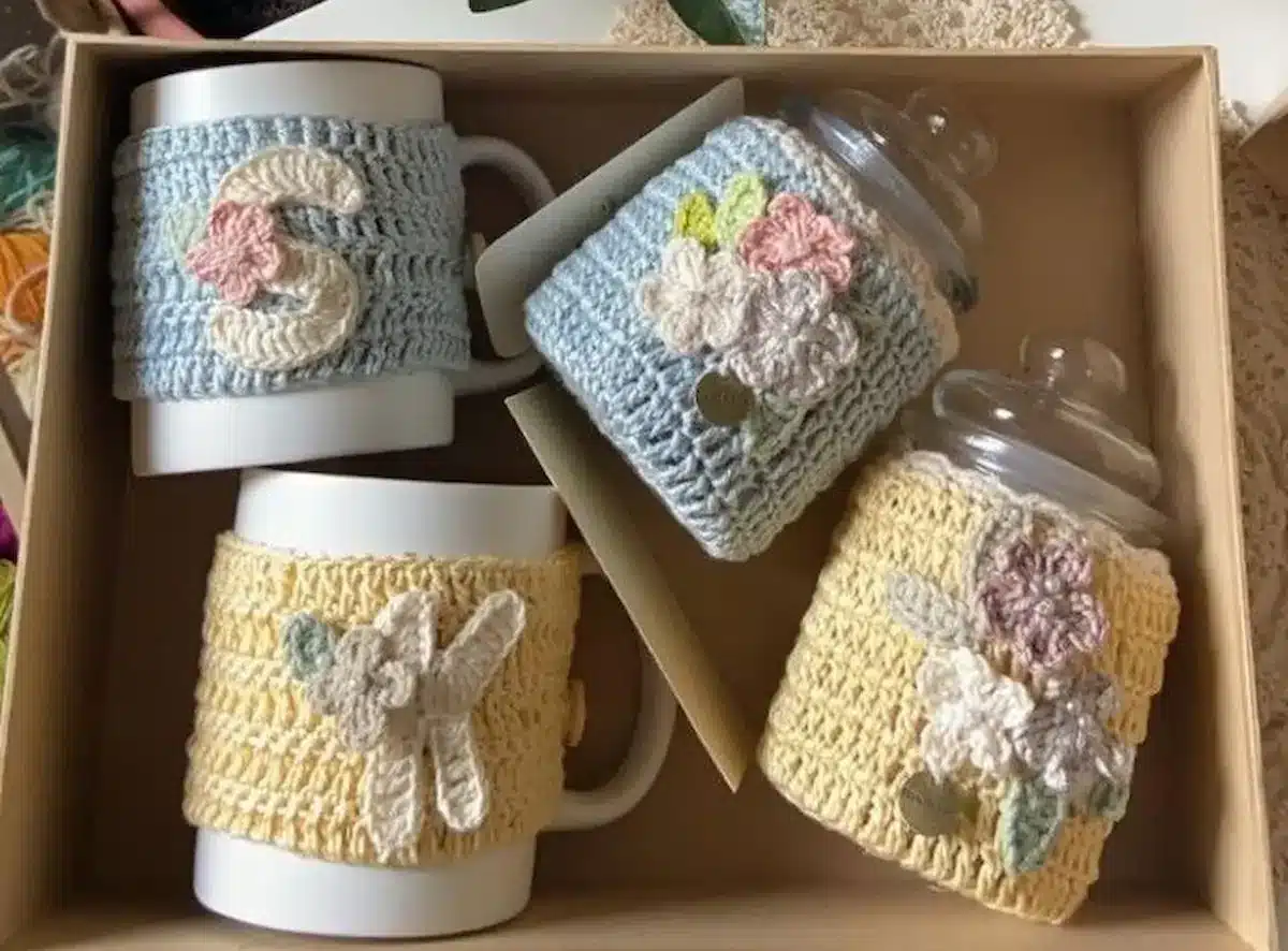 Copos de café com capas de crochê coloridas, com detalhes florais e iniciais, em caixa de presente.