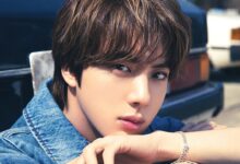 Foto do cantor Jin do BTS, com cabelo castanho, vestindo jaqueta jeans e usando uma pulseira prateada, com um fundo desfocado de um carro.