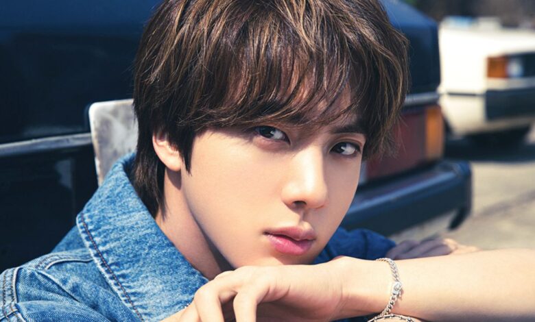 Foto do cantor Jin do BTS, com cabelo castanho, vestindo jaqueta jeans e usando uma pulseira prateada, com um fundo desfocado de um carro.