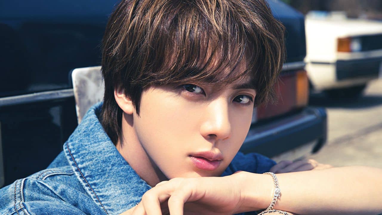 Foto do cantor Jin do BTS, com cabelo castanho, vestindo jaqueta jeans e usando uma pulseira prateada, com um fundo desfocado de um carro.