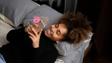 Jovem sorrindo enquanto assiste algo no celular, com ícones de interações sociais ao fundo. Imagem para mensagem de boa noite para WhatsApp.