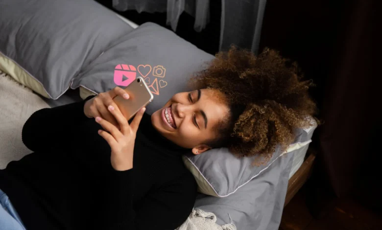 Jovem sorrindo enquanto assiste algo no celular, com ícones de interações sociais ao fundo. Imagem para mensagem de boa noite para WhatsApp.