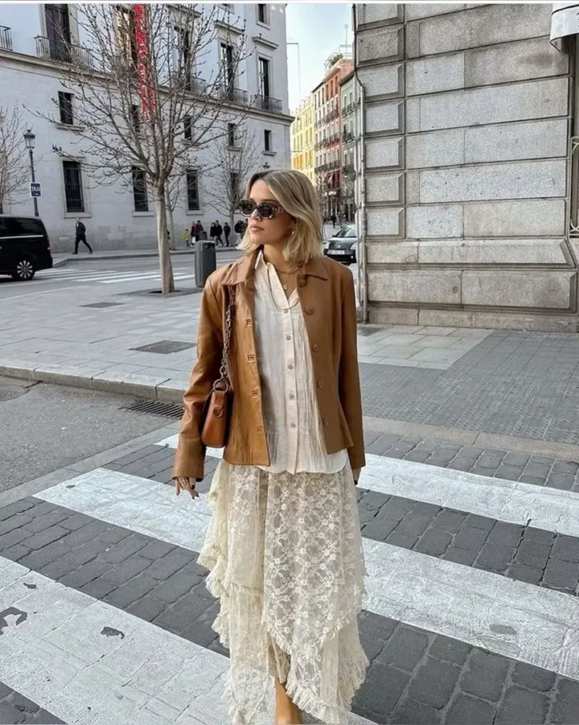 Mulher com jaqueta de couro e saia assimétrica de renda, cruzando a rua em uma cidade, com óculos escuros e bolsa de ombro.
