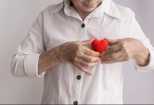 Mulher de meia-idade segurando um coração de crochê vermelho próximo ao peito, simbolizando cuidado e saúde cardíaca. Ideal para artigos sobre prevenção de infarto.