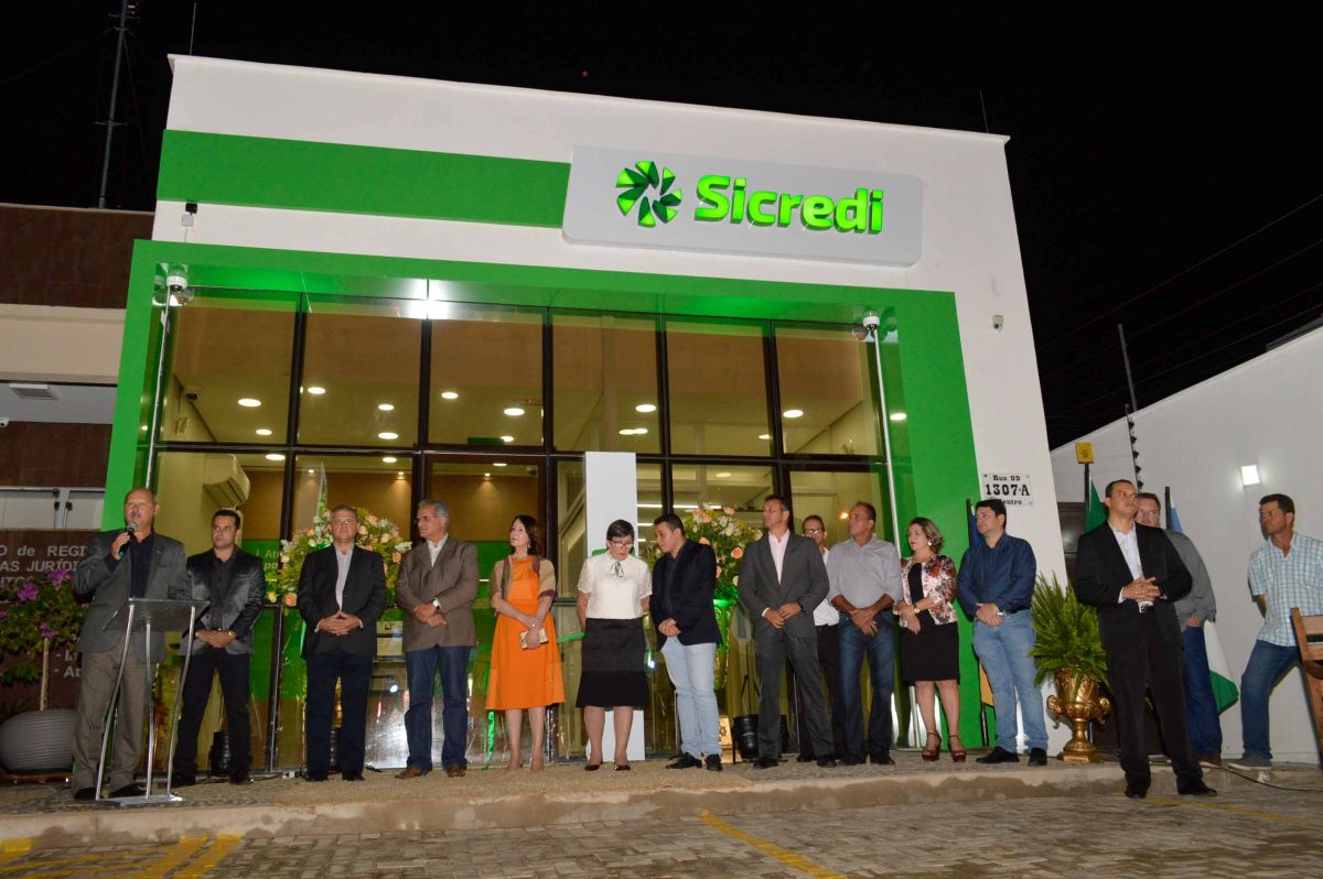  Grupo de pessoas com executivos do Sicredi posam em frente a uma agência iluminada à noite.